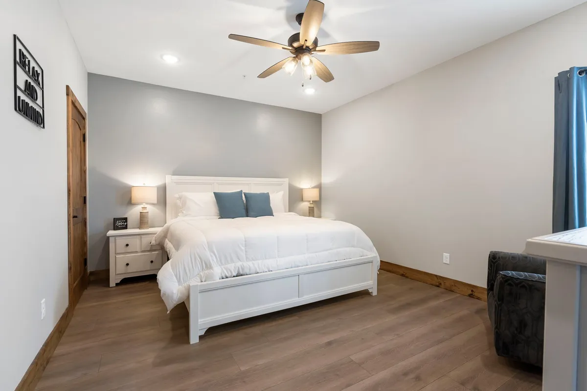 A tranquil bedroom featuring a bed with a white comforter and blue pillows, a nightstand with a lamp, a window with blue curtains, and calming wall art that says 'Relax and Unwind.' Natural light fills the room, creating a restful atmosphere.