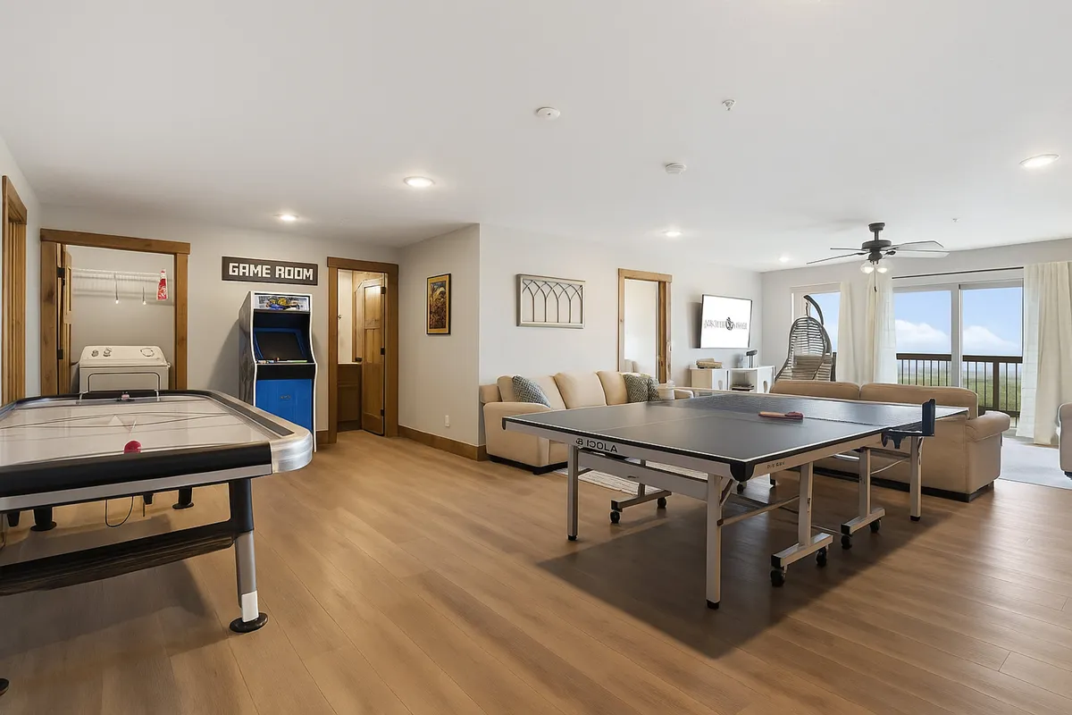 A vibrant game room featuring a ping pong table, air hockey table, arcade game machine, and a comfortable seating area. Large windows let in natural light and offer a view of the outdoors, creating a lively yet relaxed atmosphere perfect for group entertainment.