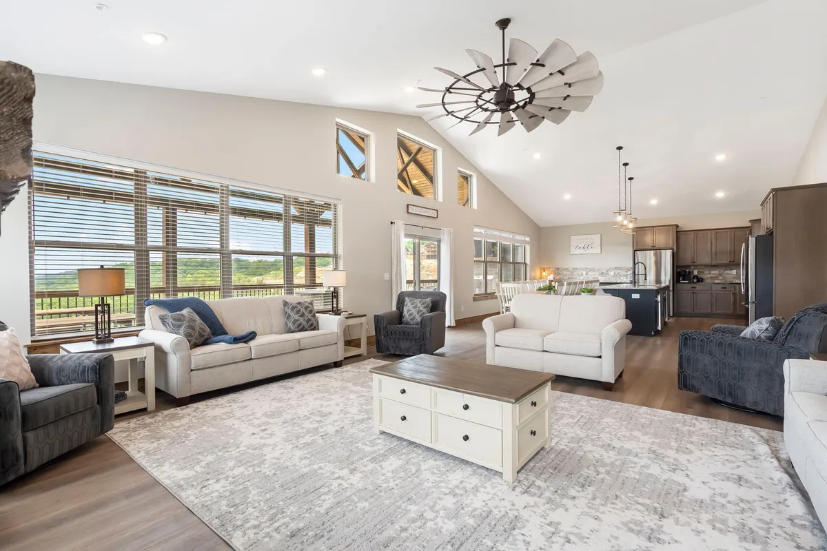 A spacious living room featuring comfortable sofas and a coffee table, with large windows that allow natural light to pour in and provide views of the outdoors. The room has a ceiling fan and is designed for relaxation and gatherings, offering a welcoming atmosphere.