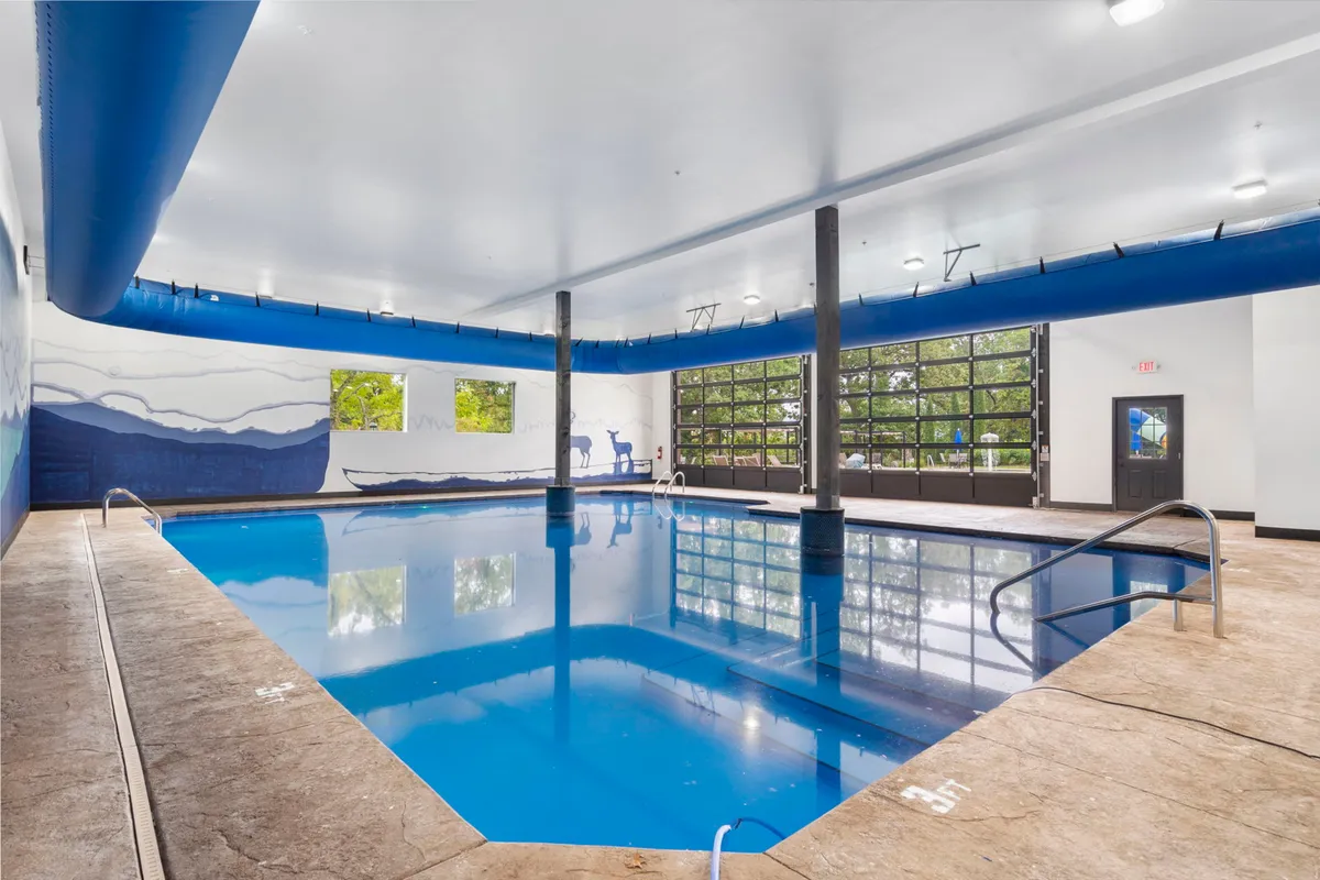An inviting interior shot of the indoor swimming pool, featuring large windows that let in natural light and a striking mountain mural along the wall. The tranquil space includes poolside seating for relaxation.