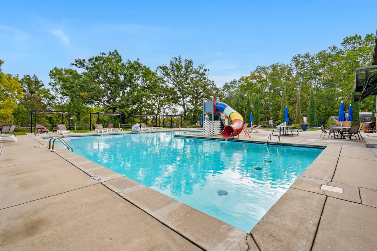 A vibrant exterior scene of the resort's outdoor swimming pool featuring an exciting water slide, plenty of lounge chairs, and tables with umbrellas for relaxation. The area is bustling with the promise of fun under the sun, perfect for families and groups.