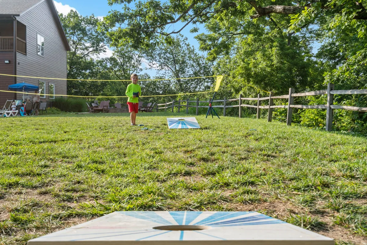 A joyful exterior image capturing a child in a bright green shirt playing cornhole near a volleyball net on a well-manicured lawn. The scene is lively and energetic, inviting guests to envision friendly competitions and active outdoor fun.