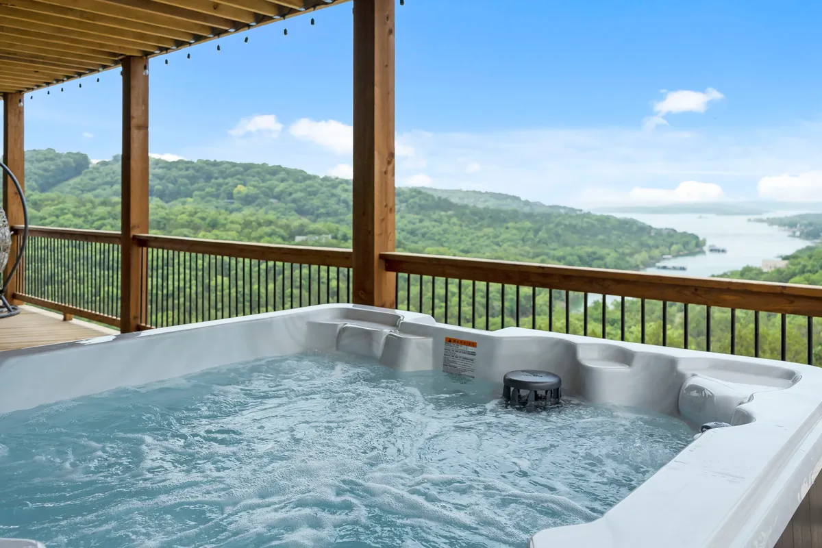 The image showcases the spacious wooden deck of a large single-family house overlooking a serene lake and rolling hills at sunset. The inviting hot tub is the centerpiece, with comfortable outdoor seating arranged for socializing, all set against a backdrop of tranquil natural beauty.